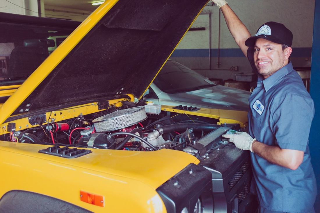 Smiling mechanic in a blue uniform next to a car with the hood up